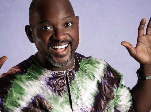 A man smiling with hands raised against a light purple background.